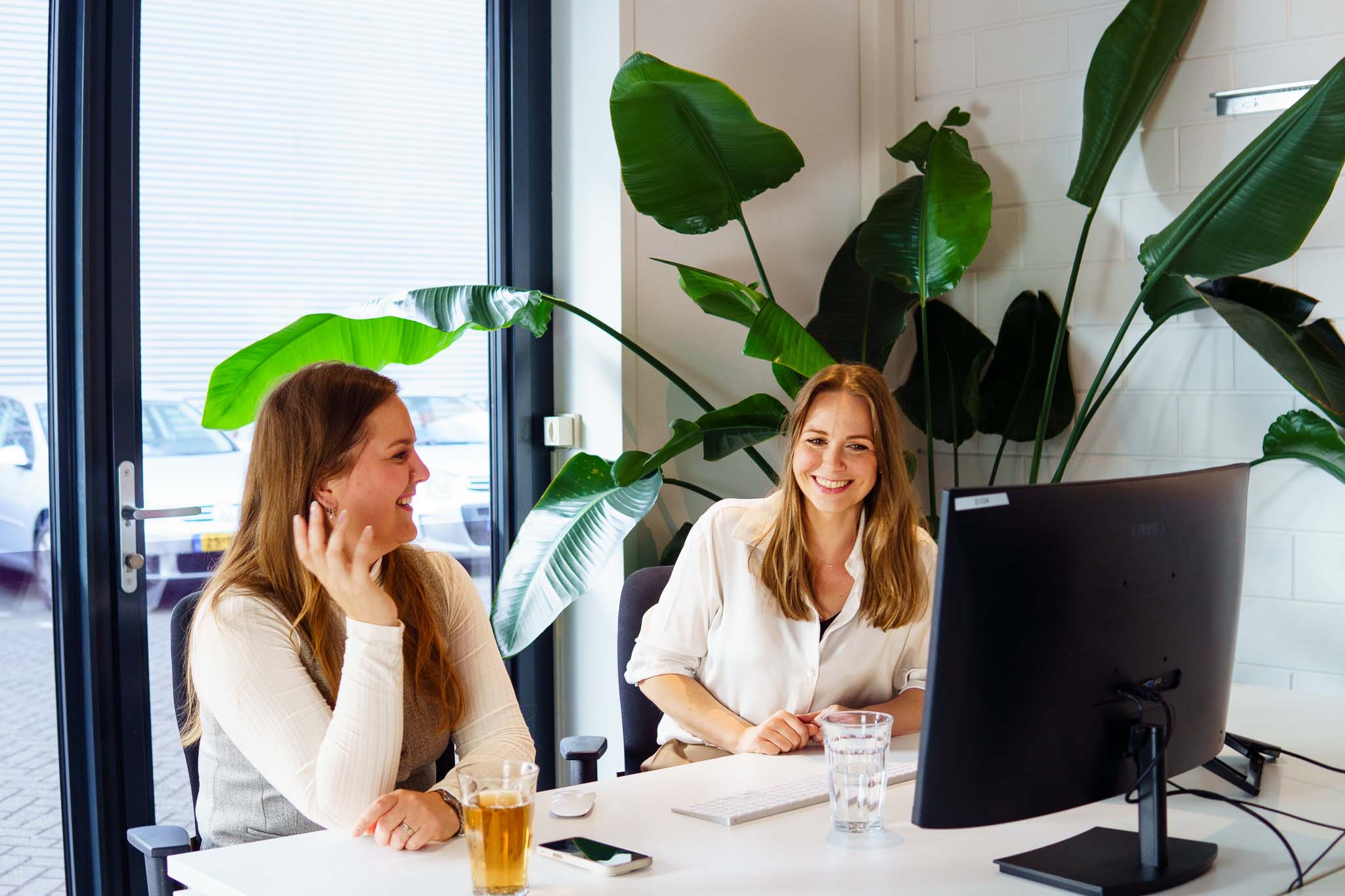 Twee vrouwen zitten lachend achter een bureau in een lichte kantoorruimte met grote planten en een computerscherm voor hen.