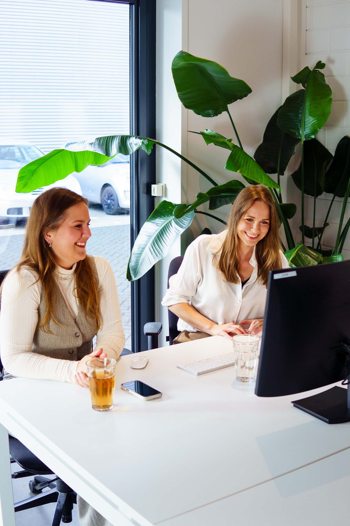 BURO210 Fullservice Marketingbureau Twee vrolijke vrouwen werken samen aan een witte bureau met een computer, drankjes en een plant in een modern kantoor. Samenwerking en productiviteit.
