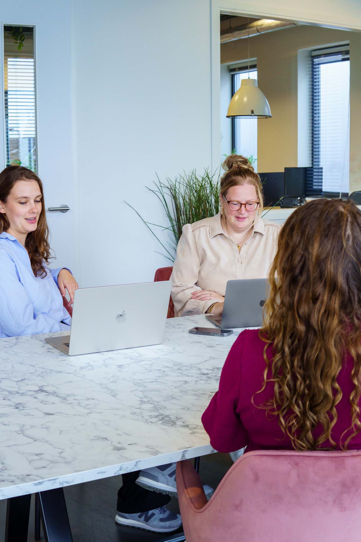 BURO210 Fullservice Marketingbureau Drie jonge vrouwen werken samen aan een marmeren tafel met laptops in een lichte kantoorruimte. Focus op teamwork en moderne werkplek.