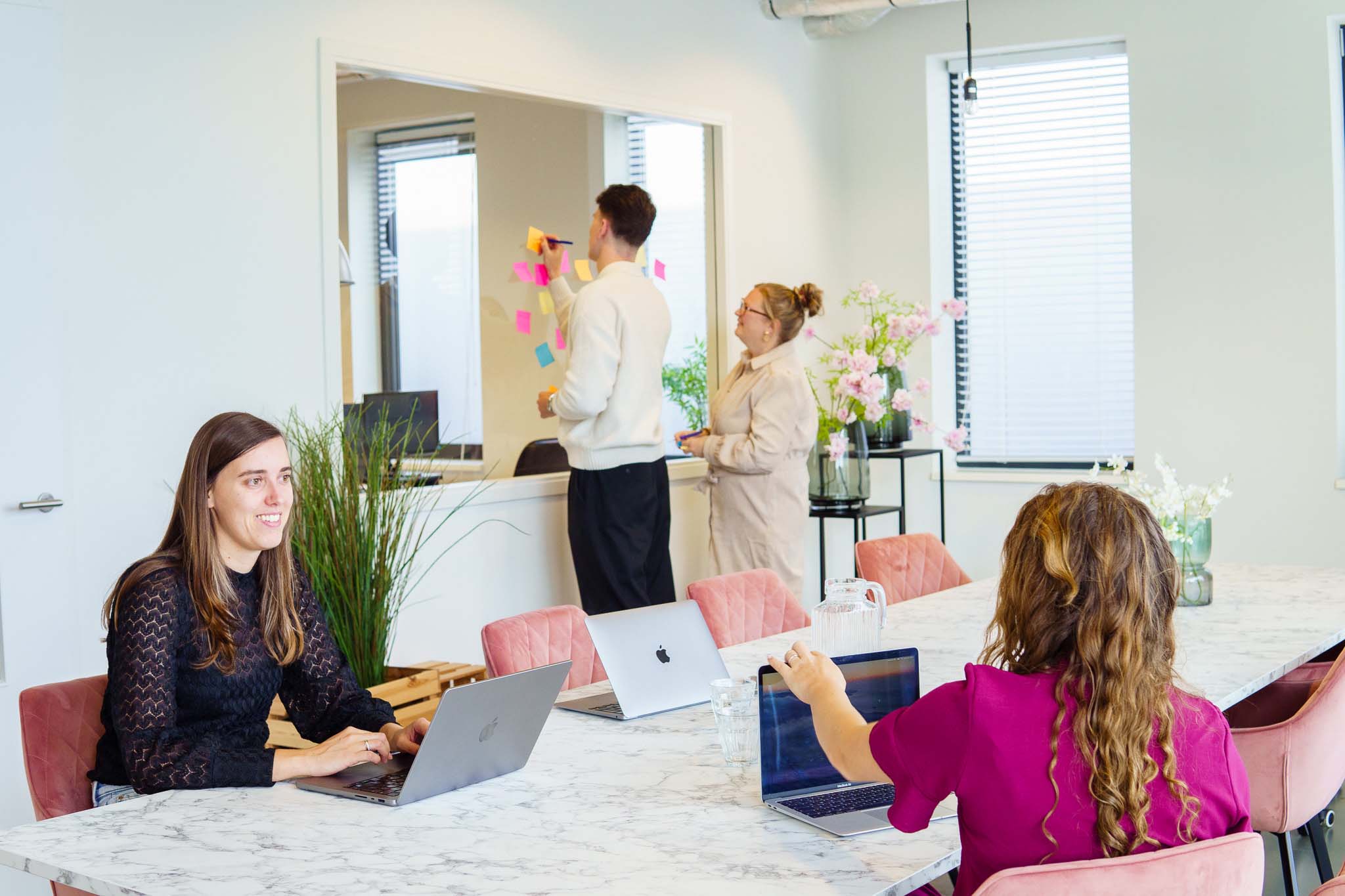 BURO210 Fullservice Marketingbureau Professionele teamsamenwerking in een moderne kantoorruimte: mensen werken met laptops aan een marmeren tafel en brainstormen bij een glazen wand met post-its.
