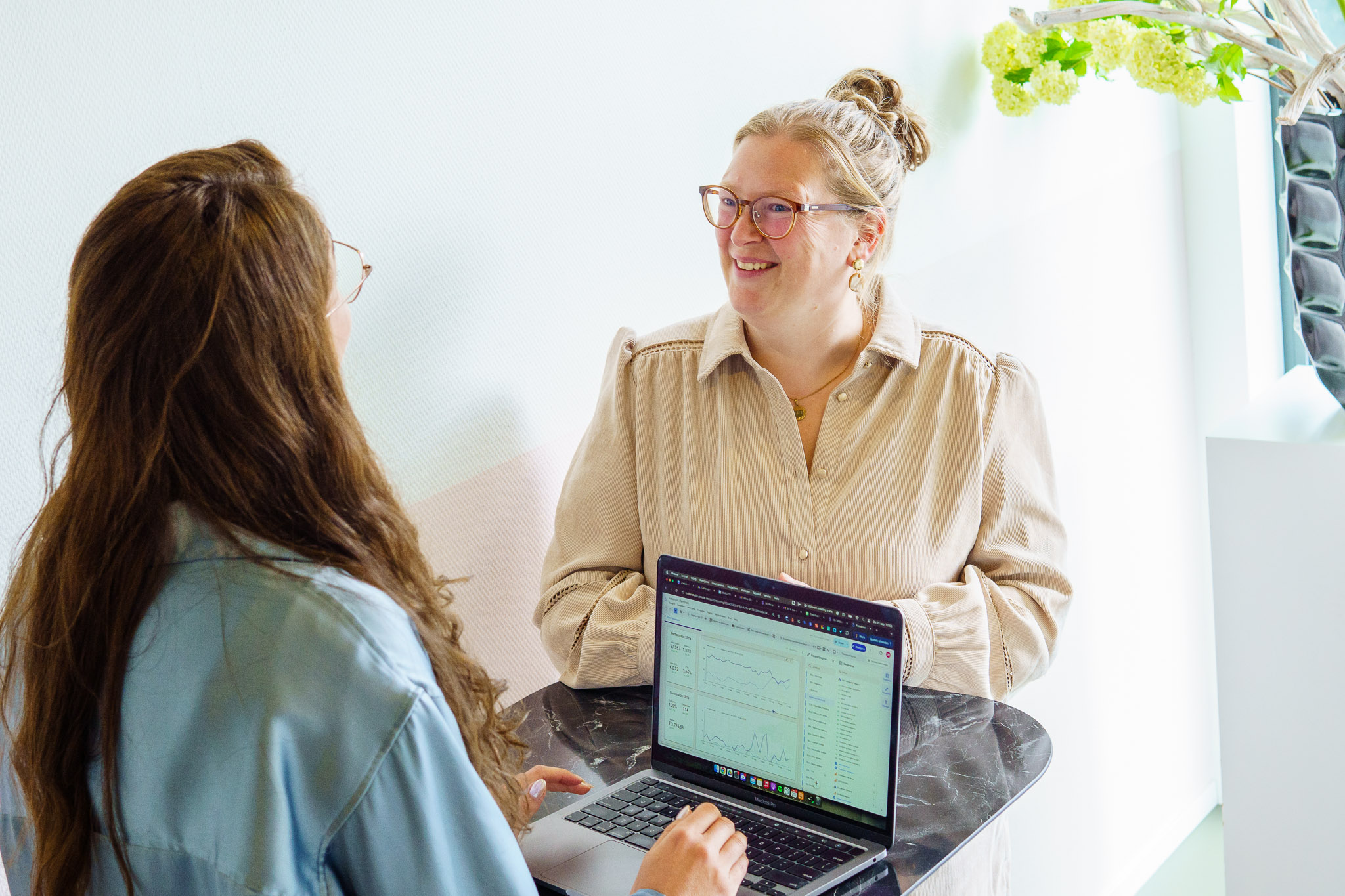 Twee vrouwen staan bij een hoge tafel terwijl een laptop met grafieken en statistieken openstaat en ze in gesprek zijn.