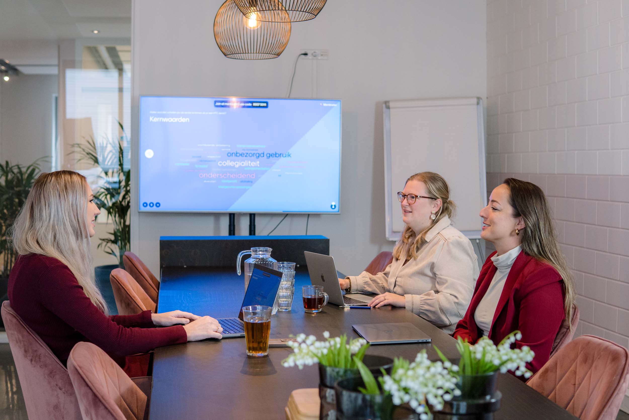 BURO210 Fullservice Marketingbureau Drie zakenvrouwen in een moderne vergaderruimte, werkend aan laptops met een presentatie over bedrijfskernwaarden op een groot scherm achter hen. Collega's samen aan het werk.
