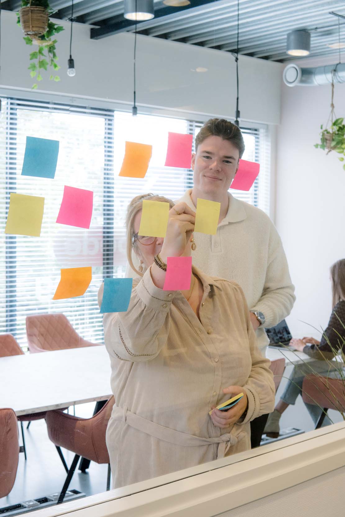 Twee personen plakken kleurrijke post-its op een glazen wand tijdens een brainstormsessie in een kantoorruimte.