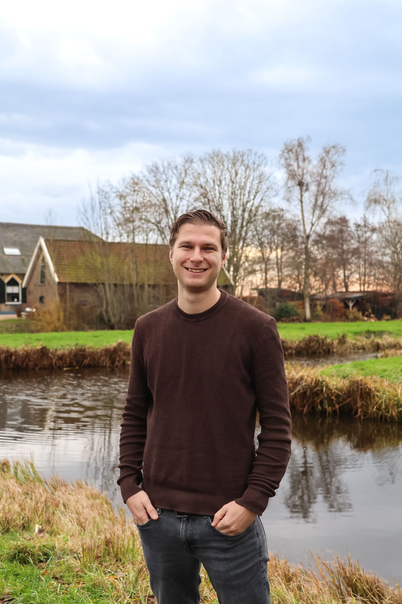 BURO210 Fullservice Marketingbureau Portret van een lachende man in bruine trui, staand bij een sloot met een traditionele boerderij op de achtergrond in een Nederlands landschap.
