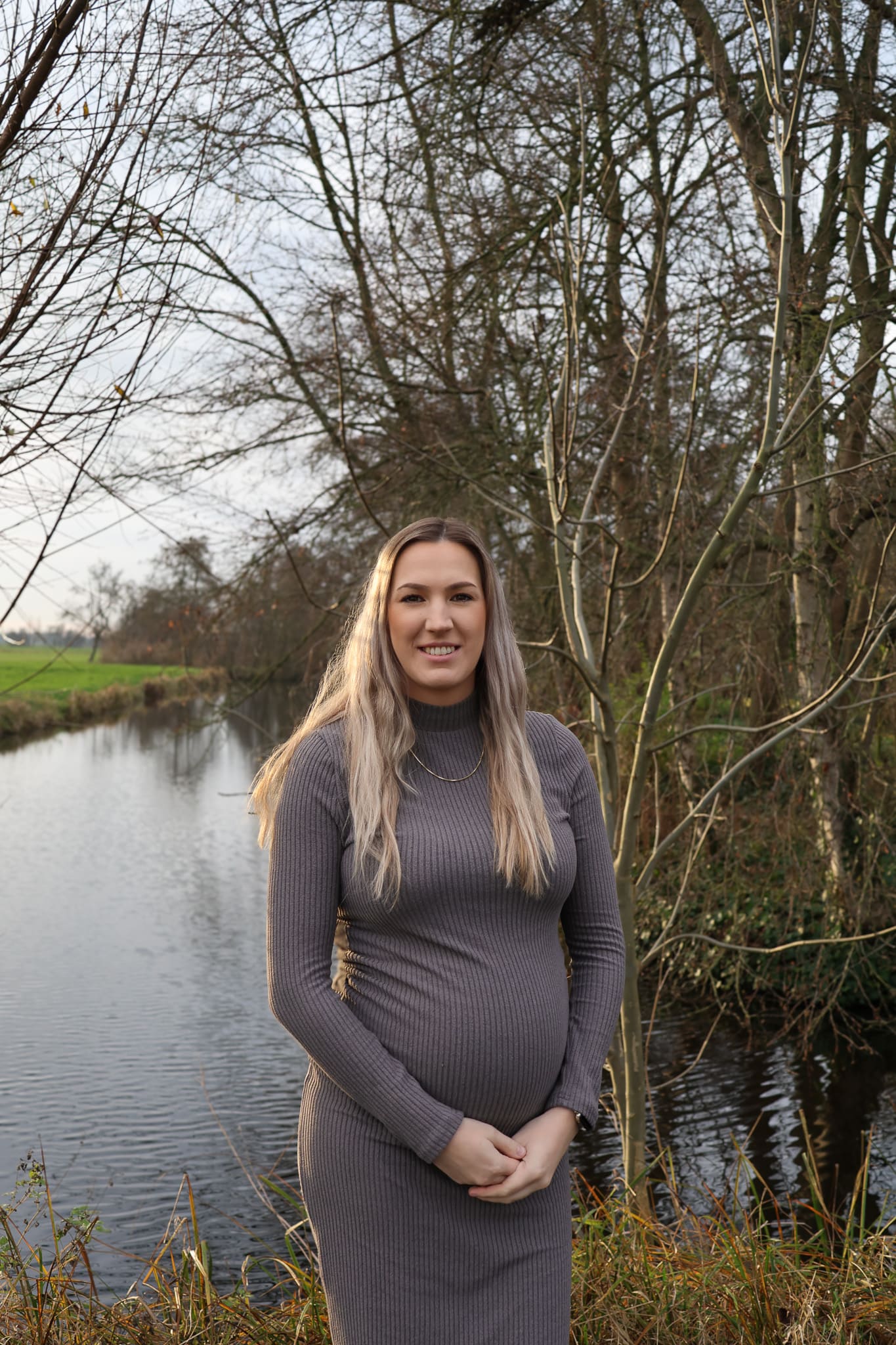BURO210 Fullservice Marketingbureau Zwangerschapsfoto van een stralende jonge vrouw met blond haar in een grijze jurk, poserend bij een rustig kanaal in de natuur. Focus op de groeiende buik.